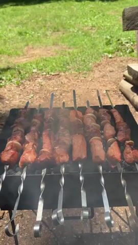 A high-angle outdoor shot shows meat skewers cooking on a grill in a grassy field. The skewers are evenly spaced and lined up on the grill. The meat pieces are brownish red, and some white onion slices are visible. Smoke rises from the grill. The background features a vibrant green grassy area, with a dirt path visible on the left. The sun is shining, and the overall scene depicts a pleasant outdoor cooking setting.