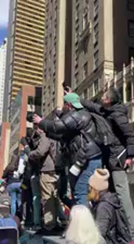 The video captures a dynamic scene of a large gathering or protest in a bustling city environment during the day. The camera starts by focusing on a group of individuals standing on an elevated platform or structure, with tall city skyscrapers dominating the background under a clear blue sky.

At the beginning (0:00-0:03), a man in a black jacket and a light green baseball cap is prominently featured. He is animated, pointing forward with his right hand and has a camera around his neck, suggesting he might be documenting the event or leading the crowd. Another person next to him also appears engaged. The crowd below them is dense and appears to be listening or participating. A voice can be heard chanting or speaking, with phrases like "...the nation... to the straight and narrow..." audible.

As the camera pans down and to the right (0:04-0:06), more of the diverse crowd becomes visible. Many individuals are present, some wearing winter attire like jackets and beanies. A woman in a purple beanie with a patterned face covering looks directly towards the camera. The street is packed with people, and colorful banners and signs in shades of yellow, green, and orange begin to emerge from the background. The tall buildings still frame the scene, but the focus shifts more to the activity on the ground.

By the end of the clip (0:07-0:09), the camera is primarily focused on the sea of people and their various banners. While specific text on the signs is mostly illegible due to movement and distance, glimpses of what appears to be "NO" and "UNION" can be seen on some banners. The overall impression is one of an energetic and engaged crowd participating in a public demonstration or parade, surrounded by the towering architecture of a major city.