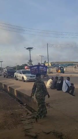 The video captures a street scene with several people and moving vehicles. A woman wearing a patterned dress carries a blue plastic basket on her head, walking towards the intersection. A group of three men are seated on a raised concrete surface next to the road, one of them holding a mobile phone. Vehicles, including a silver sedan and a motorcycle, pass through the intersection. Overhead, power lines crisscross, suspended by utility poles. In the background, there are buildings and trees, and a solar-powered street light stands near the road. The weather appears to be overcast, with a cloudy sky.