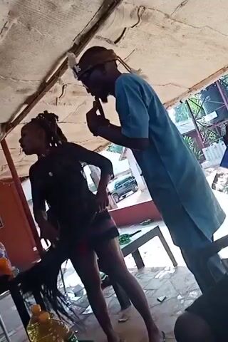 The video shows two people, a woman and a man, standing under a covered outdoor area. The woman is wearing a black top and a short skirt with stripes. She has dreadlocks and is holding a black object that looks like a chair or a piece of fabric. The man is wearing a blue outfit and a beaded necklace, along with a hat with a feather and glasses. He is standing next to her and talking, gesturing with his hands. There are tables with drinks and bottles on them. Two other men are sitting at one of the tables, one of whom is smoking. The setting appears to be a casual outdoor bar or social gathering place.