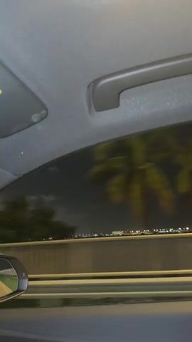 The video is a first-person perspective shot from inside a car at night. The camera is pointed upwards and outwards, showing the car's interior ceiling and a grab handle. Through the car's window, a city skyline is visible in the distance, illuminated by numerous lights of various colors. The sky is dark with some clouds. The car appears to be moving, as indicated by the slight blur of the distant city lights and the reflection of the road in the side mirror, which is partially visible on the left side of the frame. The overall ambiance is one of traveling through a city at night.