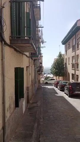 The video shows a narrow cobblestone street lined with buildings and parked cars under a clear blue sky. The buildings are multi-story structures with balconies, windows, and various architectural styles. Several cars are parked along the street, including a black Kia, a red car, and a white car. A man is seen walking in the distance. There are also trees, streetlights, and trash bins visible. The street appears to be in a town or city, possibly in Europe, given the architecture and the license plates on the cars. The camera pans and zooms in and out, providing different perspectives of the street.