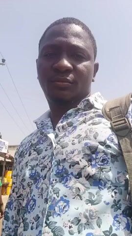 A man with short black hair is recording himself outdoors. He is wearing a light blue collared shirt with a blue and white floral pattern and has a tan backpack strap slung over his right shoulder. The background shows a street scene with buildings, signs, and utility poles. The weather appears sunny and clear. He is looking directly at the camera.