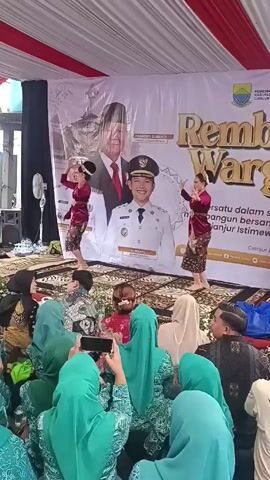 The video captures a performance of traditional Indonesian dance for an audience, likely at a local government event. Two women dressed in red tops and patterned brown skirts are dancing on a small platform decorated with rugs. They wear traditional headdresses, and their movements are coordinated and graceful.

Behind them, a large banner displays the words "Rembug Warga Cianjur Istimewa" along with photos of two men, presumably local officials. The backdrop is a combination of white fabric with the event's banner.

In the foreground, an audience is seated on the floor, watching the performance. Many of them are wearing green hijabs. Several people are holding up their phones, recording the dance. The setting appears to be an outdoor or semi-outdoor venue under a tent or awning with white and red striped material. A fan is seen in the background, providing ventilation.