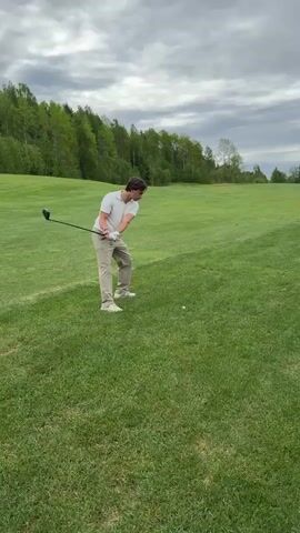 A young adult male wearing a white short-sleeved shirt and khaki pants swings a golf club on a grassy golf course. The sky is overcast. He follows through with the swing and looks towards where the ball travels, then glances to his left. A line of trees is visible in the background.
