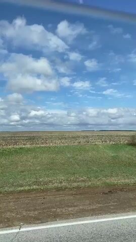 The video shows a view from a vehicle moving down a road. On the left, there is a field, part of which has short green grass and part of which looks like planted rows. The sky is blue with scattered puffy white clouds. The side mirror of the vehicle comes into view, reflecting the road and landscape.