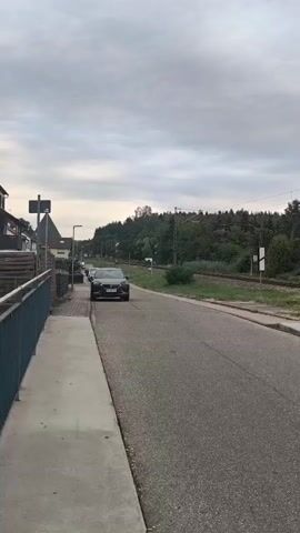The video shows a street scene with a sidewalk and a road. On the left side of the road, there is a sidewalk with a metal fence. A few houses are visible behind the fence. The road has some parked cars and a tree. In the background, there is a railway track with a train passing by. Several houses and a church tower are visible in the distance. The sky is overcast with clouds.