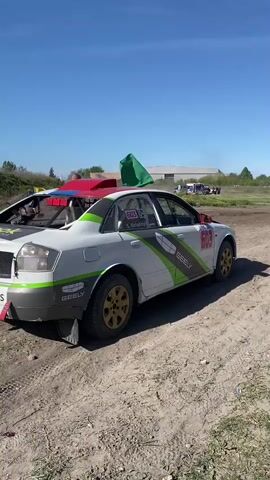 A modified white Audi rally car, marked with sponsor decals including the word “GEELY,” sits on a dirt track. The car has a roll cage, a red panel on the roof, and the rear windshield and parts of the trunk appear to be removed. The car is number 603. Someone waves a green flag to signal the car to go. The car revs its engine, kicking up dust as it drives away from the camera. The environment is outdoor, featuring other vehicles in the background, including an emergency vehicle. The sky is clear and blue.  A website address, “BELCARTEAM.RS,” is visible on the back of the car. The surface is a dirt and gravel road.