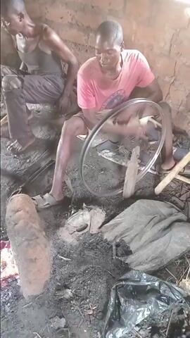 This video captures scenes from a traditional workshop in Gerkawa, Nigeria, showcasing two men engaged in manual craftsmanship. The setting is rustic, with simple, presumably mud-brick walls and a dirt floor, indicating a humble but functional workspace.

In the first scene (0:00-0:09), a man wearing a pink t-shirt and light shorts sits on the ground, operating a hand-cranked blower to stoke a fire. This blower is ingeniously fashioned from a bicycle wheel, highlighting resourcefulness and improvised technology. The fire glows intensely with red coals, suggesting it's being prepared for metalworking or blacksmithing. Another man, in a dark tank top and dark pants, is partially visible in the background, observing or waiting. The ground around them is cluttered with ashes, debris, and various tools, underscoring the nature of their work.

The video then transitions to focus on the second man (0:09-0:19, 0:22-0:30), who is engaged in woodworking. Sitting barefoot on the ground near a doorway that opens to a bright, green outdoor area, he skillfully uses a curved hand tool, possibly an adze, to chip and shape a piece of wood. The ground in this area is covered with wood shavings, indicating active carpentry. His movements are precise and rhythmic, demonstrating a practiced hand in traditional wood carving or shaping.

The scenes emphasize the manual, labor-intensive nature of these crafts, performed with focused determination. The natural lighting suggests the activities take place during the day. No explicit text is visible. The video offers a glimpse into artisanal work, where traditional methods and tools are employed to create or repair items.