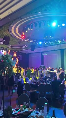 The video shows a lively indoor event, likely a wedding reception or celebration, with a large group of people dancing in a circular formation in the center of a ballroom. The room is decorated with vibrant lighting, including spotlights and colored gels, creating a festive atmosphere. Tables laden with food and drinks are visible in the foreground, with guests seated and observing the dancing. The ceiling features a circular design with an abundance of lights and a textured green surface, possibly artificial foliage. The overall scene suggests a joyous occasion filled with music and communal celebration.