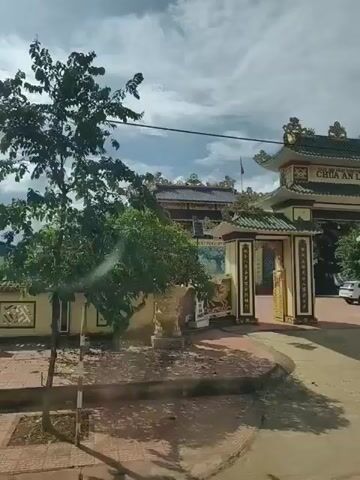 The video shows an ornate temple entrance with traditional Asian architectural elements, including tiled roofs, gold trim, and decorative carvings. The main gate has the words "CHUA AN LAC" visible at the top. On the left side of the entrance, there is a stone lion statue. Behind the gate, a building with a similar architectural style can be seen, along with trees and vegetation. A flag flies from a pole on top of the building. A car is parked to the right of the entrance. The ground in front of the temple is a dirt or gravel road. The sky is cloudy. The video appears to be filmed from a moving vehicle, as indicated by the slight camera shake and the perspective of the road. There are two figures visible in the foreground, wearing hats, suggesting they are passengers in the vehicle.