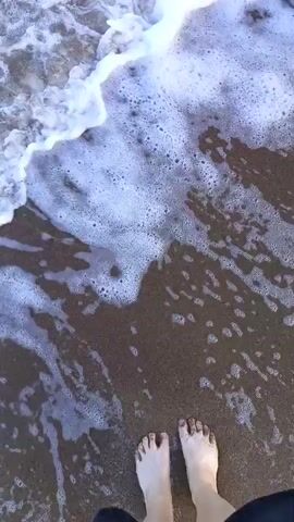 This is a first-person perspective video clip of a person standing on a sandy beach. The scene is dominated by the ocean, with waves gently washing over the person's feet. The sand appears wet and dark where the waves have receded, and foamy white water surges towards the shore. The person's bare feet, with sand clinging to them, are visible in the foreground, grounded on the wet sand. The water is clear enough to see the sand beneath its surface. The overall mood conveyed is one of tranquility and enjoyment of a natural setting. The light suggests it is daytime, possibly late afternoon or early morning given the soft quality of light. There are no visible text elements in the video. The location is identified as Yarovoye, Russia, which is known for its steppe climate and saline lakes.
