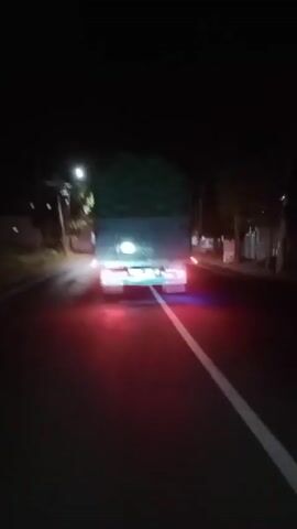 A short video clip shows a rear view of a truck at night. The truck is on a paved road with a white line dividing the lanes. The truck has its brake lights and possibly other red and blue lights illuminated. The truck appears to be loaded with green material. Streetlights and some buildings are visible on the sides of the road. The overall impression is of a vehicle driving on a road in a dimly lit area after dark.
