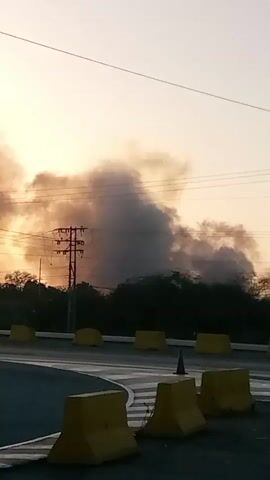 The video depicts a roadside scene at dawn or dusk, marked by a sky filled with dark, billowing smoke. This suggests a fire in the distance, as the smoke obscures the sun, casting a hazy, orange glow in the sky. Power lines and telephone poles stretch across the horizon, silhouetted against the smoky sky. In the foreground, yellow concrete barriers line the edge of the road, curving slightly to the left. The road itself is dark and wet, reflecting the ambient light. The general tone is ominous, due to the visible smoke and the dim lighting. There are no people or animals visible in the video.