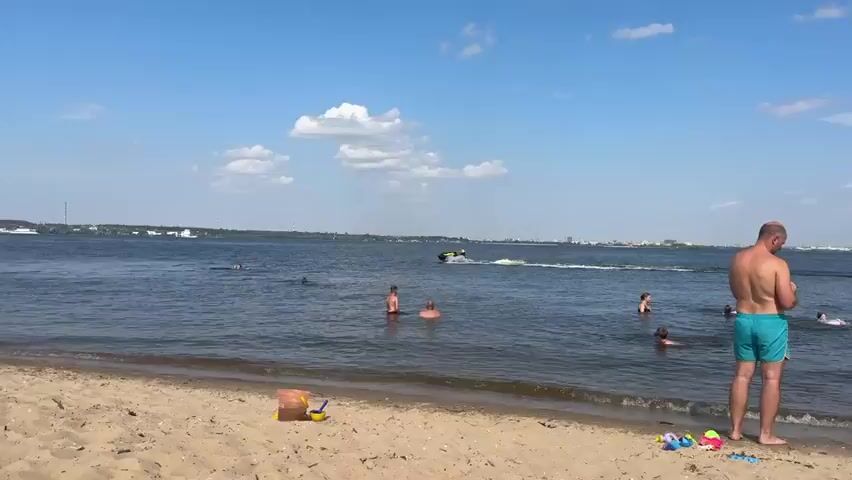 A daytime video captures a wide shot of a body of water under a bright, partially cloudy sky. In the foreground, several people are in the water, some appearing to be swimming or wading. To the right, a jet ski with a rider in a yellow vest is moving across the water, creating a white wake. In the background, a distant shoreline is visible, lined with trees and some buildings, with industrial smokestacks or towers further in the distance. The overall scene depicts a recreational activity on a sunny day at a large body of water.