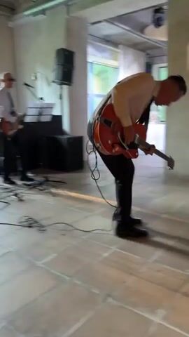 A band performs live music indoors, with one guitarist in the foreground energetically playing an orange electric guitar. He is wearing a white dress shirt and black tie, black pants, and black shoes. He is leaning forward and looking down at his guitar as he plays. Behind him, another band member, wearing a white dress shirt, black tie, and sunglasses, plays a guitar, and is partially obscured by the foreground performer. In the background, tables are set with white tablecloths and what appear to be water bottles, suggesting a dining or event setting. The room has exposed concrete ceilings and industrial-style lighting.