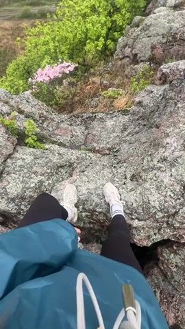 The video shows a person standing on a rocky outcrop, then pans out to show the landscape. The person is wearing a blue jacket, black leggings, and white sneakers. They are standing on gray rocks with lichen. Green trees and pink flowers are growing nearby. The camera pans up to show a wider view of the landscape, including a large body of water in the distance. The camera then shows a person with a red baseball cap and tan jacket standing on the rocks, looking out at the view. The landscape is mostly flat, with patches of grass and trees. The sky is gray and overcast.