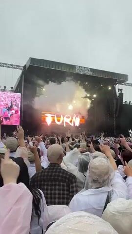 The video captures a live outdoor concert. A large stage with a massive screen is visible, displaying flashing lights and text. Smoke effects are being used on stage, creating a dramatic atmosphere. A large crowd of people is gathered in front of the stage, many with their hands raised in the air. The audience appears to be enjoying the performance, with some filming the event on their phones. The sky is overcast, suggesting potential rain, and some attendees are wearing clear plastic ponchos. The overall scene is one of excitement and energy, typical of a large music festival.