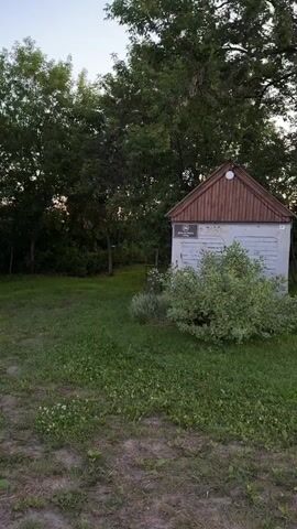 The video begins with a blurry shot of a small, gray structure with a brown, gabled roof, surrounded by green grass and trees. A white sign is affixed to the structure. The camera then pans to the right, capturing a view of trees and a clear blue sky. The scene transitions to a dirt path alongside lush green foliage, with power lines visible in the distance. The video concludes with a view of a tranquil pond reflecting the pastel colors of the sunset sky, with a tall wooden power pole standing nearby and trees bordering the horizon. The overall impression is a serene, rural landscape at dusk.