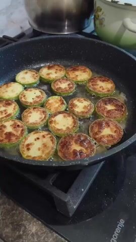 In a close-up shot, slices of zucchini are being fried in a black non-stick pan. The zucchini slices are arranged in a single layer, overlapping slightly, and appear to be golden brown and slightly crispy. The pan is on a black glass stovetop with "Gorenje" text visible at the bottom right. In the background, out of focus, are two pots, one metallic and the other white with a floral pattern. The scene appears to be indoors, in a kitchen, possibly during the daytime, given the lighting. The activity is cooking, specifically frying zucchini. There are no people visible in the frame, and no discernible emotions are conveyed. The setting suggests a home kitchen environment in Sokolivske, Ukraine.
