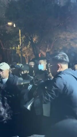 The video shows a lively scene with a crowd gathered around musicians playing instruments in an outdoor setting, seemingly at night. The musicians include a man in a suit playing two small trumpets simultaneously, and a man in a white t-shirt enthusiastically playing a drum. Many members of the crowd are watching and smiling, while others are wearing masks. Most attendees are older Asian males wearing hats. In the background, trees and a street light can be seen, indicating a park or urban environment. The atmosphere appears festive and celebratory.