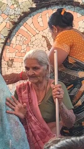 An elderly woman with gray hair, wearing a green blouse, a pink and red shawl, and a beaded necklace, is standing and holding a silver cane with both hands. She is surrounded by other people and looks slightly to the left. A woman in a yellow polka-dot top is behind her. Behind them is a wall covered in colorful tiles. The woman is standing outdoors.