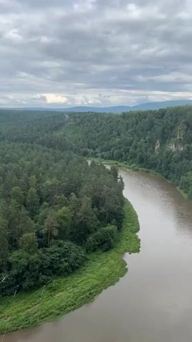 This is a vertical, aerial shot of a wide, meandering river flowing through a lush, green, forested valley under a cloudy sky. The river, with its murky brown water, takes up the right half of the frame, curving from the distance towards the foreground. The left bank of the river is lined with dense green foliage, including trees and grasses. The surrounding hills are covered in a thick, dark green forest of coniferous trees, with some deciduous trees interspersed, creating a rich, textured landscape.

In the distance, the valley recedes, with layers of hills fading into a hazy horizon under the overcast sky. The clouds are a mix of grey and white, with patches of brighter sky peeking through, suggesting a day with variable weather, possibly damp or humid, with a hint of mist or fog clinging to the distant cliffs on the right side of the river. The overall mood is serene and natural, highlighting the raw beauty of the wilderness.

On the lower left of the frame, there's a hint of human activity. A few thin, dark cables stretch diagonally across the green bank, possibly part of a zip line or some other recreational apparatus. This suggests that the location is a place where people might visit for outdoor activities.

There is no visible text in the media. The time of day appears to be daytime, given the overall illumination, but the cloudy conditions obscure any direct sunlight or specific shadows that would pinpoint the hour. The location cues, such as the type of forest and the terrain, combined with the provided context of Kul'metovo, Russia, suggest a natural, possibly mountainous or hilly region within that area. The scene evokes a sense of vastness and natural splendor, undisturbed by excessive human presence, save for the subtle suggestion of recreational infrastructure.