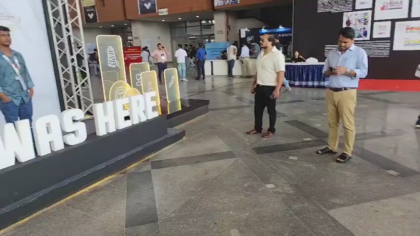 The video opens with a wide shot of an indoor exhibition hall. In the foreground, a large, three-dimensional art installation spells out "I WAS HERE" in white block letters. To the left of the installation, a young man in a blue shirt and jeans stands facing forward, looking towards the camera. Behind him, a banner advertises the "25th Dhaka International Textile & Garment Machinery Exposition." To the right of the "I WAS HERE" installation, a man in a light-colored polo shirt and dark pants stands facing away from the camera.

The video pans slowly to the left, revealing more of the exhibition hall. Several other people are visible in the background, walking and standing around various booths and displays. The floor is a patterned grey tile. The scene is well-lit and appears to be a busy event. The video continues to pan and zoom out, showing more of the hall and the people attending the exhibition. The overall atmosphere is one of a bustling trade show or conference.