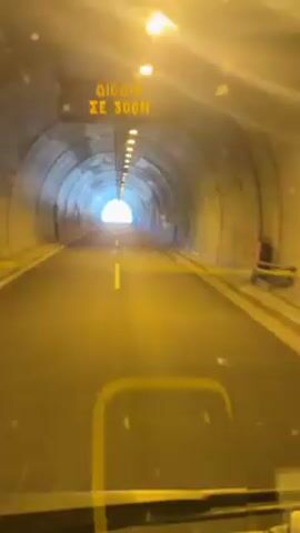 A forward-facing, shaky, first-person perspective shot shows a vehicle moving through a tunnel. The tunnel has a concrete, arched ceiling with a row of evenly spaced, small, yellow lights running down the center. The road is asphalt with a solid yellow line dividing it in the middle. The end of the tunnel shows a bright, white light.