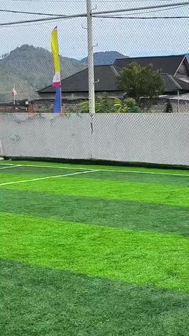 A group of people are playing soccer on an artificial turf field with white markings. The field is enclosed by a net fence. In the background, there are mountains and buildings. One person in a yellow shirt is near a soccer goal. Other people are running and kicking a soccer ball. The weather appears to be overcast.