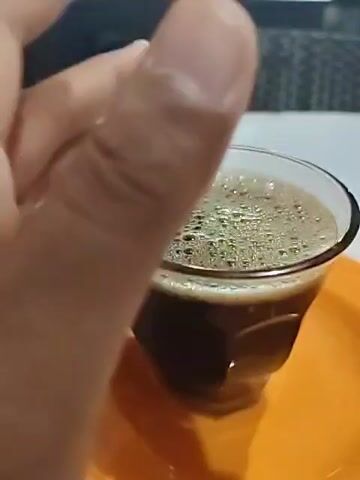 Here's a detailed description of the video:

The video shows a person stirring a cup of dark coffee with a silver spoon. The coffee is in a clear, ribbed glass sitting on a small orange plate. The coffee has a foamy top with lots of tiny bubbles. The person stirs the coffee, then leaves the spoon in the glass. The background is slightly blurred but seems to show an outdoor setting, possibly a cafe or restaurant, with tables and chairs visible. There are other people sitting in the background. The table in the foreground is white. The lighting suggests it might be evening or nighttime.