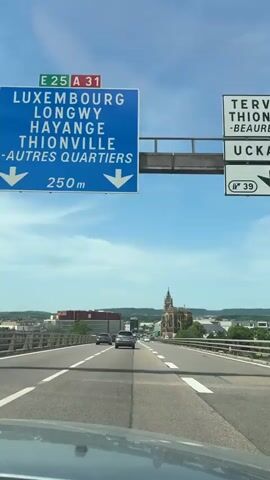 The video depicts a view from inside a car, driving along a highway, with a cityscape visible in the distance. The sky is blue with scattered clouds. The road has multiple lanes, and other cars are present. A prominent building resembling a church stands out in the cityscape. A speed limit sign of 70 is visible. The camera is static, and there are no apparent interactions.
