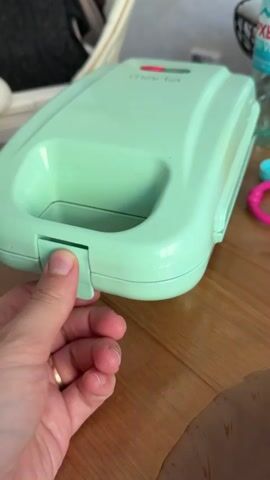 A hand opens a small, light-green waffle maker on a wood table. Inside the waffle maker is a golden-brown waffle. A bottle of water and some colorful plastic toys are nearby on the table.