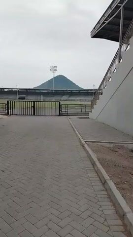 The video shows a gray brick-paved walkway leading towards a large stadium with black metal gates.  In the background, a small green hill rises behind the stadium. A tall light tower stands atop the hill. To the left of the walkway are stairs leading up to the stadium.  Two people in red shirts are standing on the stairs. A person in a white shirt is bent over near the bottom of the stairs. To the right of the walkway are stairs leading down, covered by a roof. The sky is overcast. Music is playing in the background.