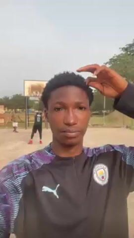 Here's a description of the video:
The video shows a young man taking a selfie video at an outdoor basketball court. He is wearing a black, long-sleeved shirt with purple and blue accents on the sleeves, a Puma logo, and a Manchester City emblem. He runs his fingers through his short, curly hair as he holds the camera. Behind him, other people can be seen playing basketball on a court with a simple hoop. The background includes trees, buildings, and a fence. The ground appears to be dirt. The weather looks bright but overcast.