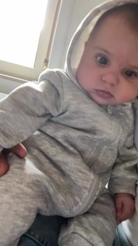 A baby in a hooded grey jumpsuit is being held in someone’s arms. The baby has a serious expression and is looking at the camera. The background includes a window and part of the person’s white shirt. At the start of the clip, a hand touches the baby’s chest area briefly.