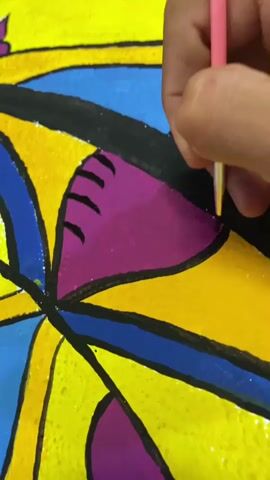 A close-up shot shows a person's hand holding a pink paintbrush and painting a colorful abstract design. The design features bold geometric shapes in yellow, blue, and purple, outlined in black. The person is meticulously filling in a purple section with the paintbrush. The texture of the painting surface appears to be rough, possibly canvas or textured paper. The lighting is bright, highlighting the vibrant colors of the artwork and the detail of the brushstrokes.