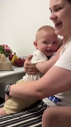 A woman is sitting and holding a baby on her lap. The baby is smiling and looking at the woman. In the background, there is a table with fruit and a basket of tissues. Another child approaches and interacts with the baby. The woman is wearing a white t-shirt and striped pants. The baby is wearing a light-colored shirt and shorts. The other child is wearing a white t-shirt. The scene appears to be indoors, possibly in a home.
