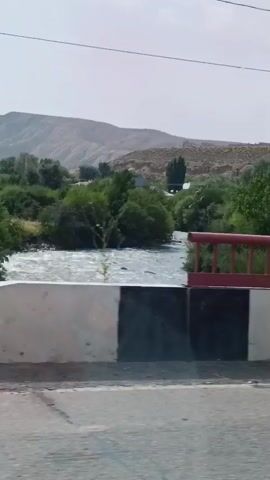 The video captures a moving shot from a vehicle traveling on a road. In the foreground, the concrete barrier of the road is visible, with a section of red railing extending over a body of water. The water appears to be flowing, with some ripples on its surface. Beyond the railing, lush green vegetation, including trees and bushes, dominates the landscape. In the background, rolling hills or mountains are discernible under a clear sky. The shot also includes utility poles and wires, suggesting the presence of infrastructure. The overall impression is of a scenic drive through a rural or semi-rural area with natural elements and man-made structures.