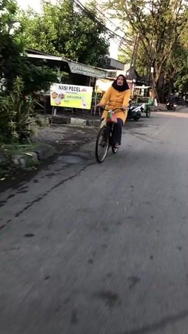 A woman wearing a yellow tunic and headscarf rides a bicycle down a paved road. She is in Gelam, East Java. The road appears wet, indicating recent rain or damp conditions. The woman is looking forward as she pedals, and her expression is neutral.

To the left of the road, there is a row of buildings that appear to be shops or food stalls. A yellow banner with Indonesian text is visible, with the words "NASI PECEL" and "NASI CAMPUR" prominently displayed. "Nasi Pecel" is a traditional Javanese dish of blanched vegetables served with peanut sauce, and "Nasi Campur" means mixed rice. Other buildings and trees are visible in the background, suggesting a moderately populated area.

Several motorcycles are parked on the side of the road, and a person can be seen near one of them. The lighting suggests it is either early morning or late afternoon, with the sun casting shadows and illuminating the scene. The overall atmosphere seems to be that of a typical day in a local Indonesian town, with everyday activities taking place. The woman on the bicycle is the central focus of the image, her journey down the road suggesting movement and purpose.
