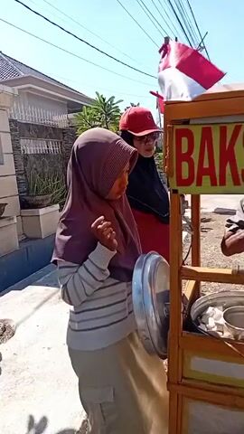 Two people are standing next to a food cart. A woman wearing a hijab and a grey sweater with stripes is looking at the cart, and her right hand is raised. Behind her, a person wearing a red cap is also looking at the cart. The food cart is made of wood and has a sign that says "BAKSO RAHAYU JOWO KHAS MALANG". The cart contains bottles of sauce and containers of food, including noodles and crackers. The scene is outdoors, with a clear blue sky and trees in the background.