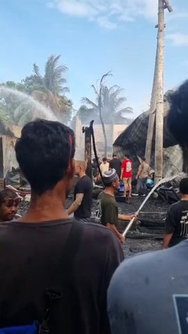 The video depicts the aftermath of a fire, with charred remains of structures visible against a partially cloudy sky. Several individuals are present, some wearing casual clothing, and others with hoses spraying water on the debris. One person is holding a hose. The environment includes palm trees, overhead power lines, and remnants of walls and roofing. The focus is on containing the fire or cooling down the area.