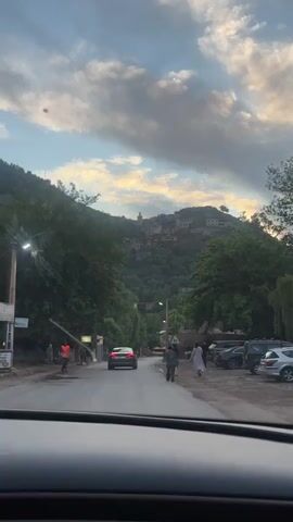The video shows a car driving down a road in a small town. There are people walking on the sidewalks. In the distance, on top of a hill, there's a village with a tower. The sky is cloudy. Trees line the side of the road. There are several parked cars on the right. A hotel sign is visible. A black car is driving in front.