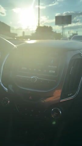 The video shows the interior of a car, with a focus on the dashboard and infotainment system. The sun is shining brightly through the windshield, causing lens flare and obscuring some details. The infotainment screen displays a Bluetooth connection. Outside the car, trees and a billboard are visible, along with what appears to be traffic. There are no people visible in the frame. The video is static, with no discernible movement or action.