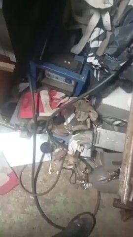 The video shows a floor in a cluttered workspace or garage. The floor is dirty and covered with various objects, including gloves, tools, equipment, and pieces of metal and plastic. There is a foot wearing a black shoe visible in the corner.