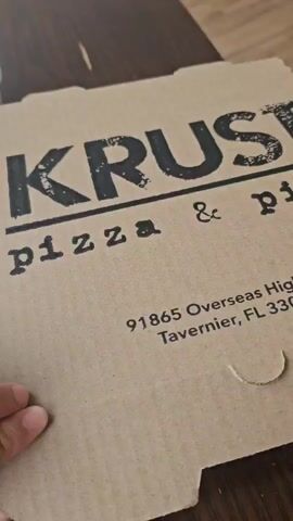 The video begins with a close-up shot of a Krust pizza box on a wooden table. The box is open, revealing a pizza inside. The pizza is square and has a tomato base, mozzarella cheese, and fresh basil leaves scattered on top. The camera zooms in on the pizza to display the toppings clearly. The box also displays the address "91865 Overseas Highway Tavernier, FL 33070" and phone number "(305) 396-7248" printed on the side. A hand is visible, holding the corner of the box.
