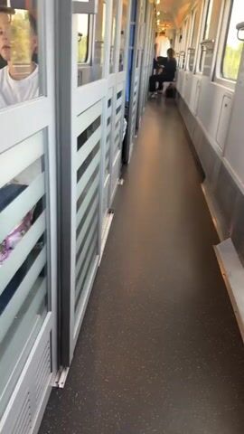 The video shows the interior of a train car, viewed from a low angle, looking down the aisle. The floor is dark gray with small white speckles. On the left side of the aisle are doors with frosted glass panels that have horizontal lines. On the right side, the wall of the train car is visible, which appears to be a light gray color. In the distance, at the end of the aisle, two people are seated on the right side of the train, facing away from the camera. The person in the foreground is wearing dark clothing, and the person behind them is also wearing dark clothing. They appear to be looking down. The lighting in the train car is moderate. The video is shaky, suggesting it was filmed while the train was in motion.