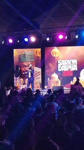 Here is a description of the video.

The video captures a live event featuring a man speaking on a stage in front of a seated audience. The speaker is wearing a suit and standing under bright stage lights. Behind him are two large screens displaying images. The left screen shows a portrait of a man in traditional attire, while the right screen features the title "SARKIN DARIYA" above a building resembling a mosque or palace with hanging lanterns. The audience, filling the foreground, is mostly obscured by darkness. Some individuals are holding up phones to record the event. The overall atmosphere suggests a comedy show or cultural event.