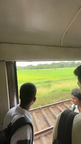 The video is shot from inside a train car looking out a window. Two young men are visible in the foreground, facing away from the camera. The man on the left, wearing a white shirt and a dark vest, looks towards the right. The man on the right, with long dark hair, is also looking towards the right. The train tracks are visible outside the window, and beyond them, a wide expanse of green fields stretches to a line of trees in the distance under an overcast sky. The overall impression is of travel through a rural landscape.