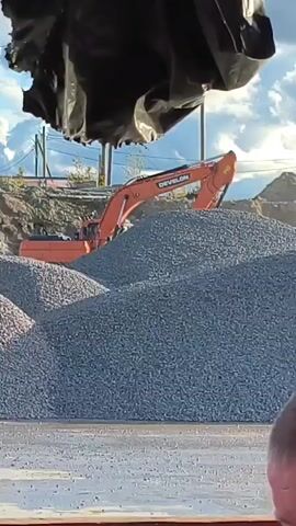 A construction site scene features a large pile of gravel in the foreground. An orange excavator with the brand name "DOOSAN" visible on its boom is partially obscured by the gravel pile, with its arm extended. In the background, there are more piles of dirt and gravel, some power lines, and a partly cloudy sky. A person's head and shoulder are visible in the bottom right corner, and another person wearing a blue jacket and a reflective vest is partially visible in the left foreground. The scene appears to be from a distance, possibly filmed through a window or opening.